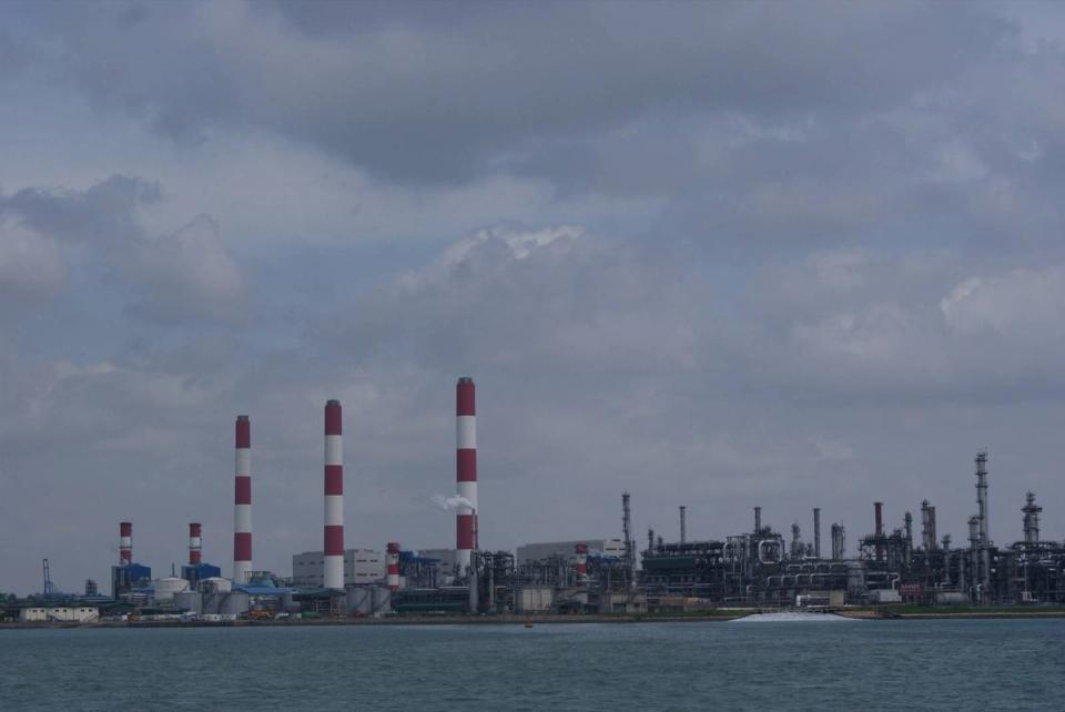 Red-and-white striped chimneys and petrochemical plant structures on Jurong Island seen across the water under a cloudy sky.