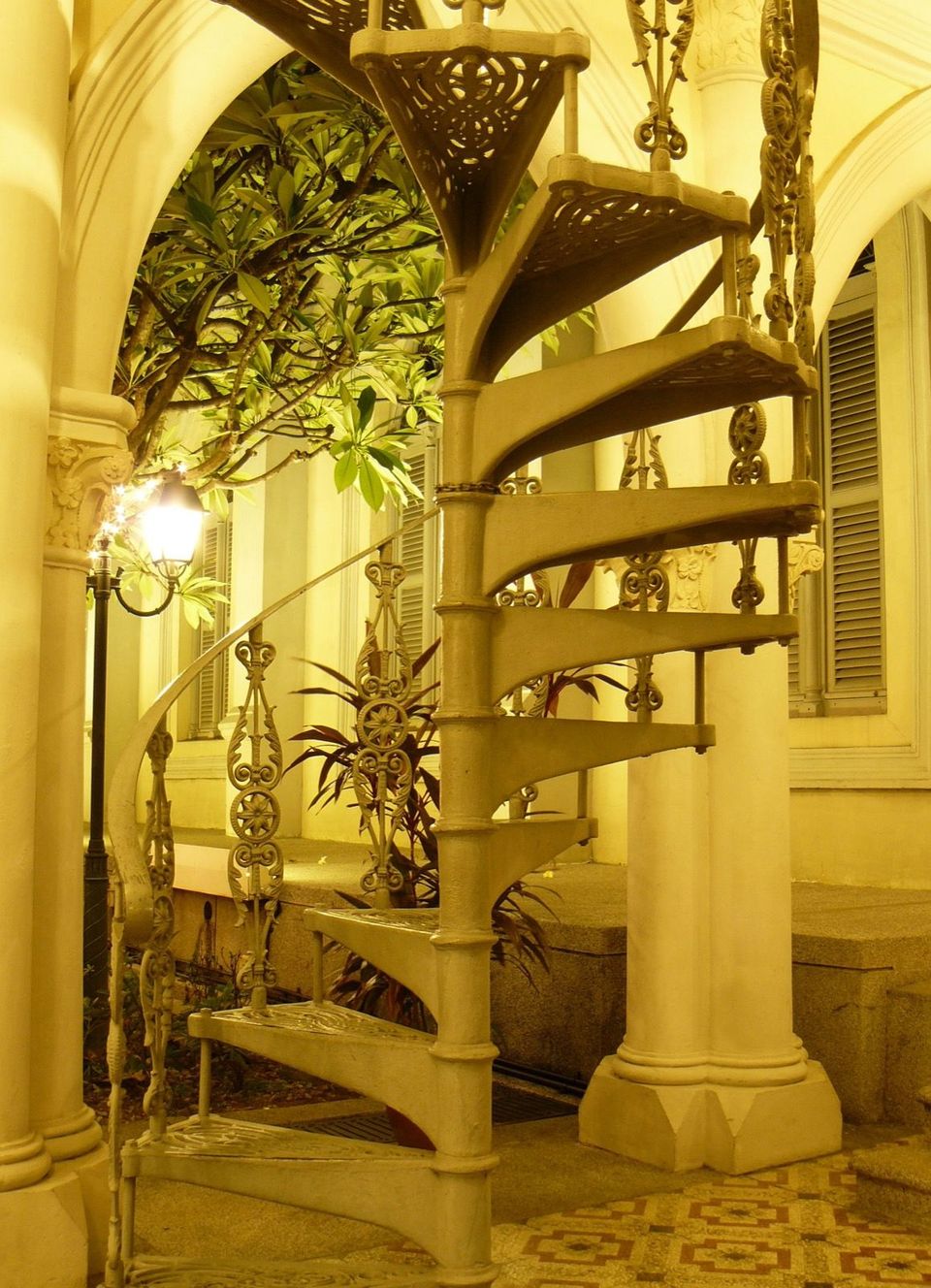 A wrought-iron spiral staircase and lamplight in a courtyard at CHIJMES, Singapore, at night.