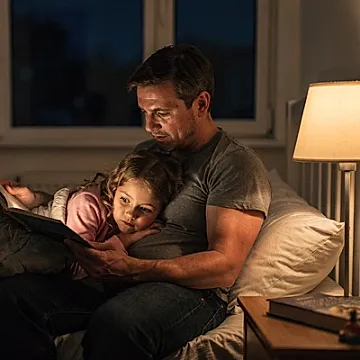 Editorial photo of a parent and young girl reading together on the edge of a bed under warm lamp light, used in an article about 5-minute bedtime stories
