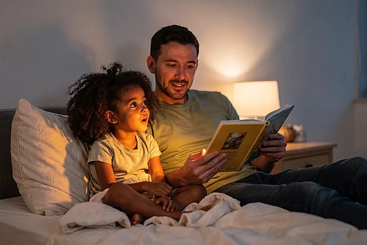 Editorial photo of a young girl turning mid-story to look up at a parent with unguarded attention during bedtime reading, used in an article about bedtime stories for kids