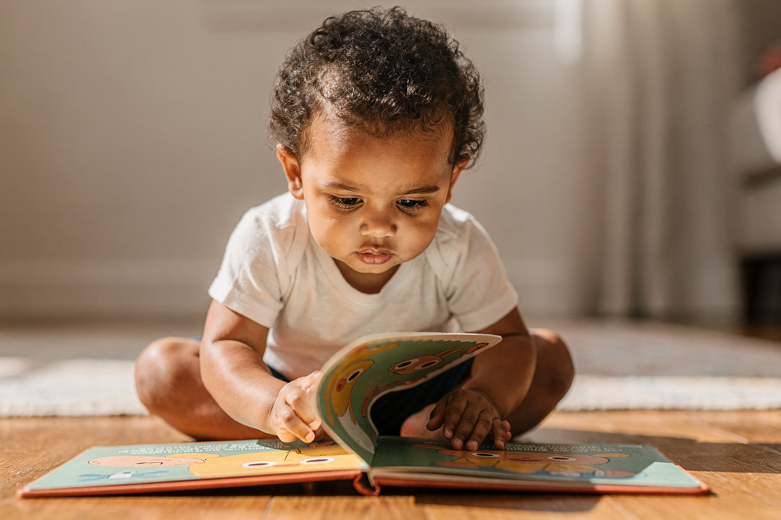 Editorial photo of a toddler with warm brown skin carefully lifting a flap in a board book, body leaning forward with concentrated anticipation, used in an article about interactive books for toddlers