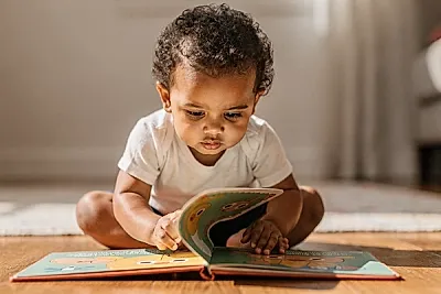 Editorial photo of a toddler with warm brown skin carefully lifting a flap in a board book, body leaning forward with concentrated anticipation, used in an article about interactive books for toddlers