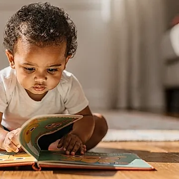 Editorial photo of a toddler with warm brown skin carefully lifting a flap in a board book, body leaning forward with concentrated anticipation, used in an article about interactive books for toddlers
