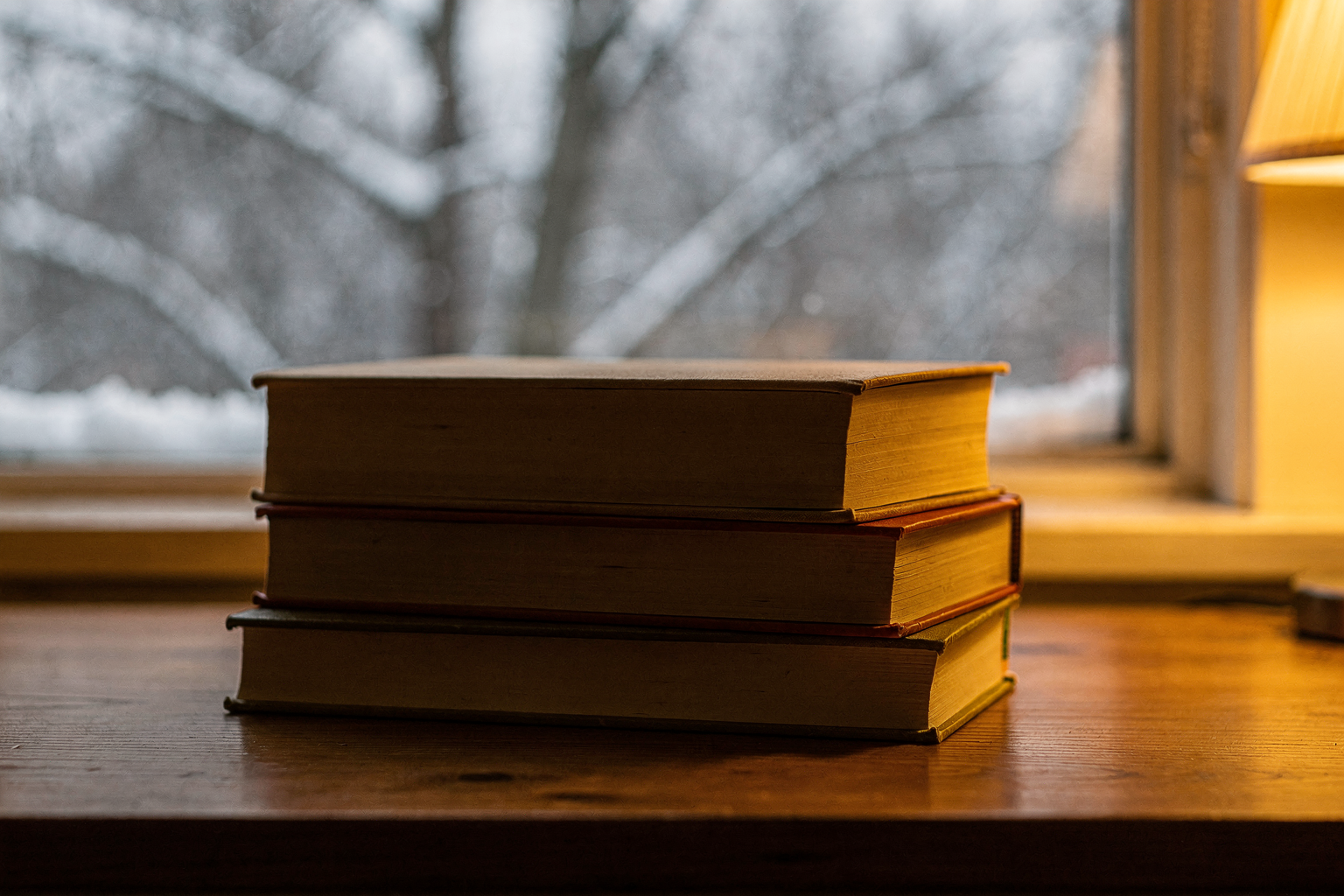 Editorial photo of a small stack of books with plain backs on a wooden table near a window with snow visible outside, lit by warm lamp light, used in an article about Christmas books for toddlers