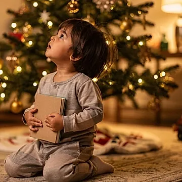 Editorial photo of a toddler boy in pajamas kneeling near a lit Christmas tree, holding a face-down book against his chest while gazing up at the lights, used in an article about Christmas books for toddlers