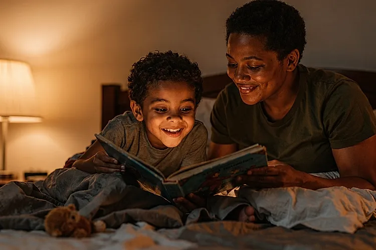 Editorial photo of a 3-year-old boy with warm brown skin chanting along to a rhyming bedtime story with eyes squeezed shut while a parent holds the book and waits, used in an article about what makes bedtime stories work for 3-year-olds