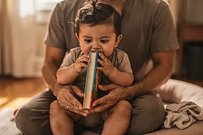 Editorial photo of a baby boy chewing the corner of a board book while a parent's hands rest nearby without intervening, used in an article about the best board books for babies