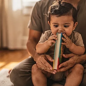 Editorial photo of a baby boy chewing the corner of a board book while a parent's hands rest nearby without intervening, used in an article about the best board books for babies