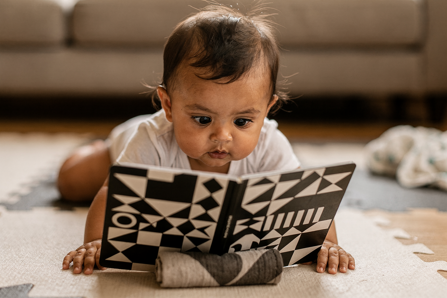 Editorial photo of a baby girl on her tummy during tummy time with her head lifted, gaze fixed on a high-contrast black-and-white board book propped in front of her, used in an article about the best board books for babies