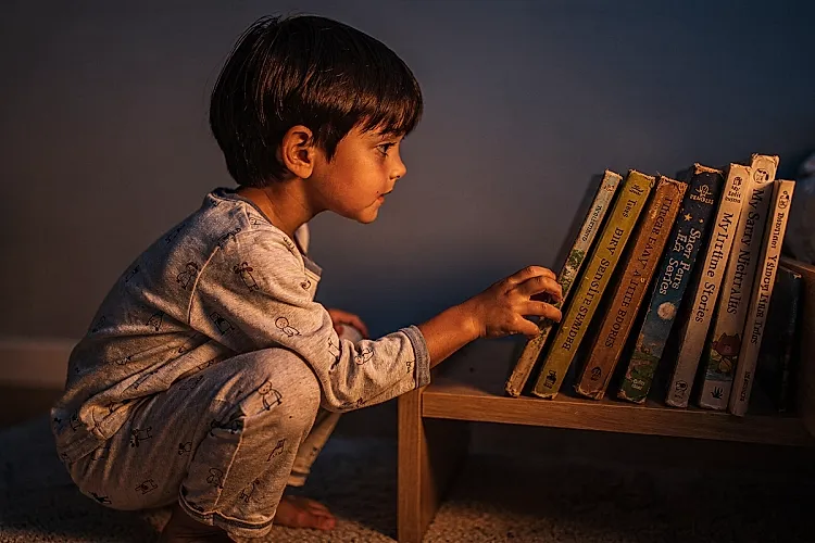 Editorial photo of a young boy with warm copper-toned skin and straight thick dark hair crouching in pajamas to choose a book from a low bedroom shelf, completely unhurried, used in an article about building a short-story bedtime routine