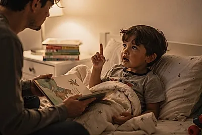 Editorial photo of a 4-year-old boy raising his hand mid-story to ask why while a parent holds an open book and pauses with quiet recognition, used in an article about bedtime stories for 4-year-olds