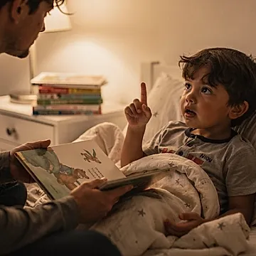 Editorial photo of a 4-year-old boy raising his hand mid-story to ask why while a parent holds an open book and pauses with quiet recognition, used in an article about bedtime stories for 4-year-olds