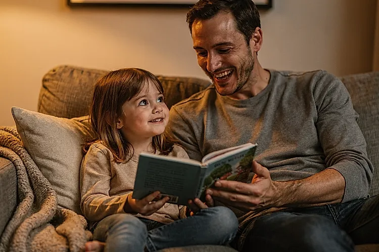 Editorial photo of a four-year-old girl looking up from a picture book with quiet certainty while her parent laughs, caught mid-correction, used in an article about reading with 4-year-olds