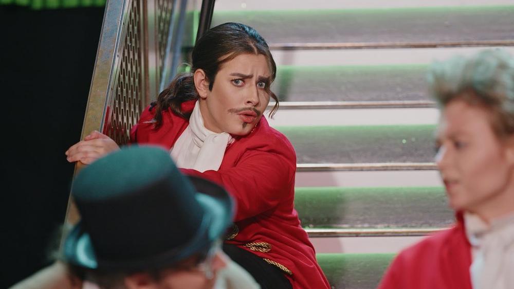 En skuespiller sitter i trappa, i rød jakke, og sniklytter på en samtale mellom en mann i flosshatt og en annen uniformert mann.