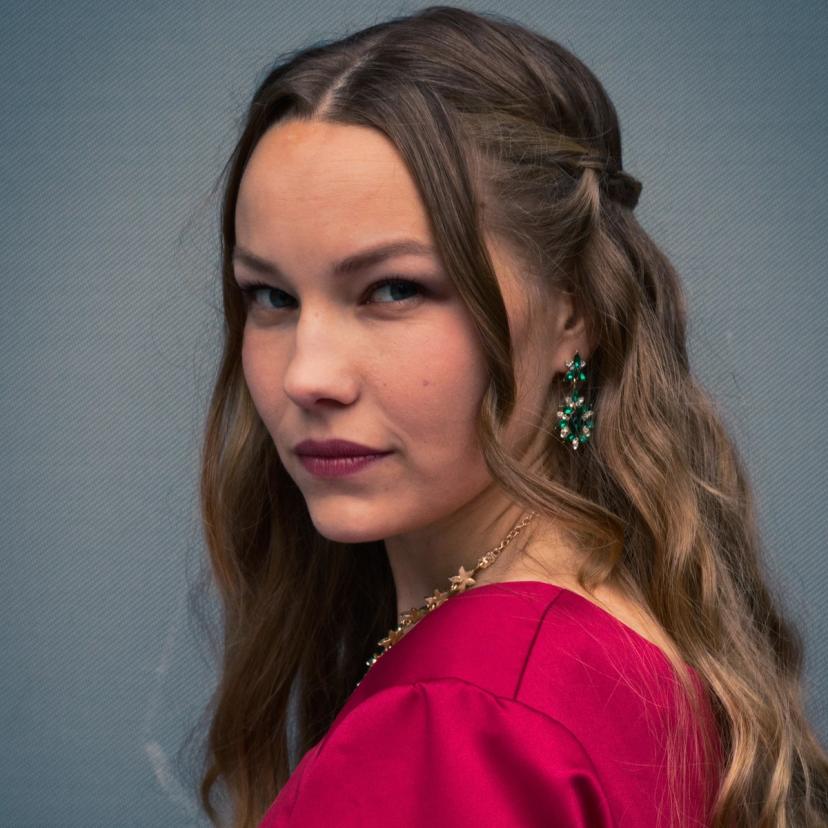 Portrettbilde av Lady Macbeth. Hun har på rødkjole, grønn øredobb, og ser inn i kamera med et lurt blikk.