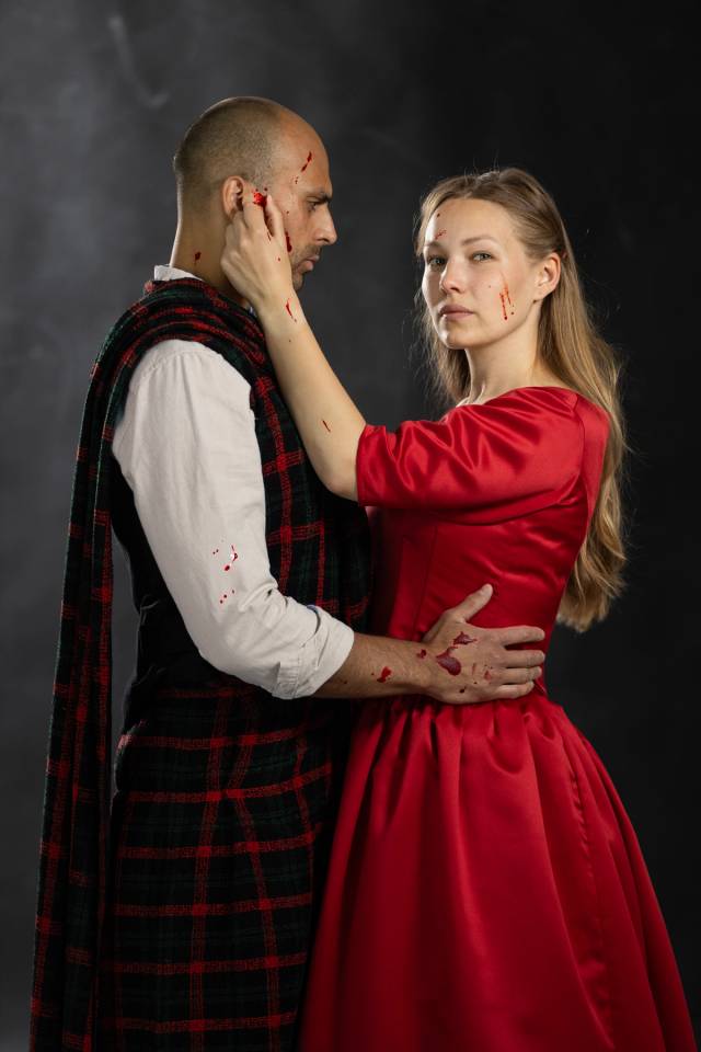 Lady Macbeth står til høyre, i rød kjole og med blod i ansiktet. Til venstre står Macbeth, i kilt, og holder armen rundt henne. Han har blod i ansiktet og på hånda.