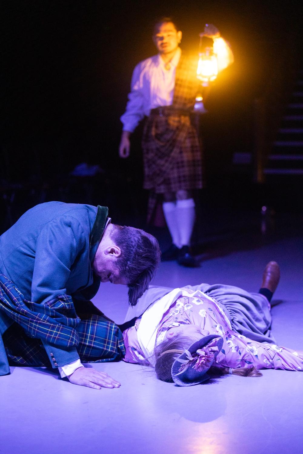 Macduff kneler på gulvet, foran den døde kona si, mens Ross står i bakgrunnen med en lykt.