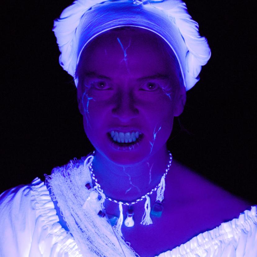 Portrettbilde av heks. Hun gløder lilla i mørket, og har lysende blodårer i ansiktet.