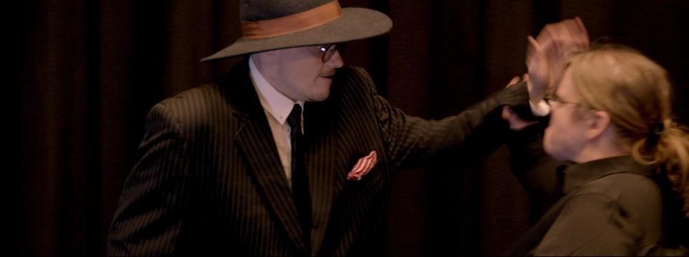 Don Leone, i hatt og dress, som prøver å slå en kvinne i svart skjorte.