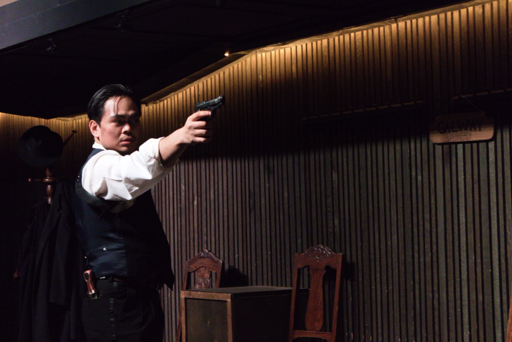 Edmund, en gangster i dress, som sikter mot høyre med en pistol. Bak er en brun vegg, brune stoler og en stumtjener det henger en svart hatt og svarte frakker på.