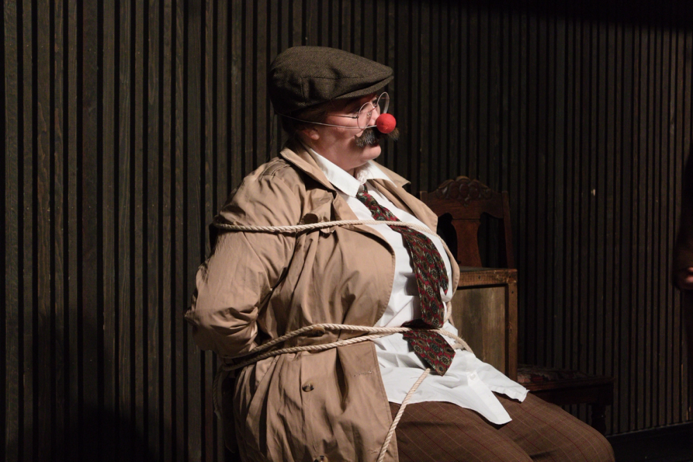 Kate er forkledd som mann, bundet til en stol, med løsbart og klovnenese.