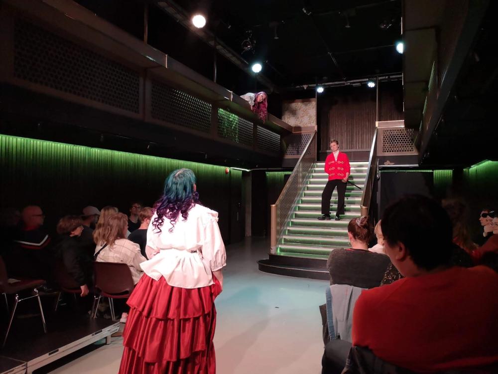 Et intimt scenelokale dominert av en trapp i midten. Oppe i trappa står en mann (spilt av en kvinne) i rød jakke og ser nonchalant ned på en kvinne i kjole som står på gulvet. Ovenfor trappa er det et lite galleri, og der titter en annen kvinne i kjole ut og sniklytter.