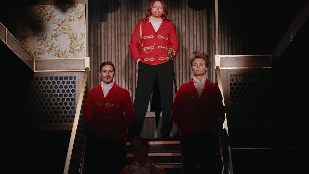 Tre skuespillere i røde uniformer. Alle er kvinner som spiller menn. De står i toppen av trappa, og det er spotlight på dem.