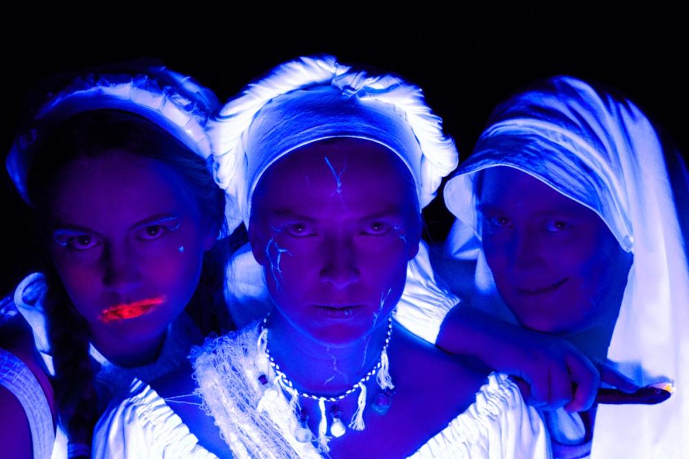 De tre heksene, i glødene hvite klær og med lilla hud.