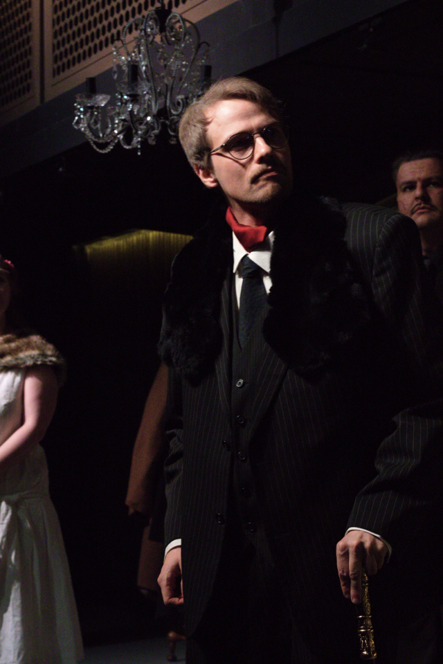 Don Leone, en aldrende mafiaboss i dress, med pels. Det henger en lysekrone i bakgrunnen.