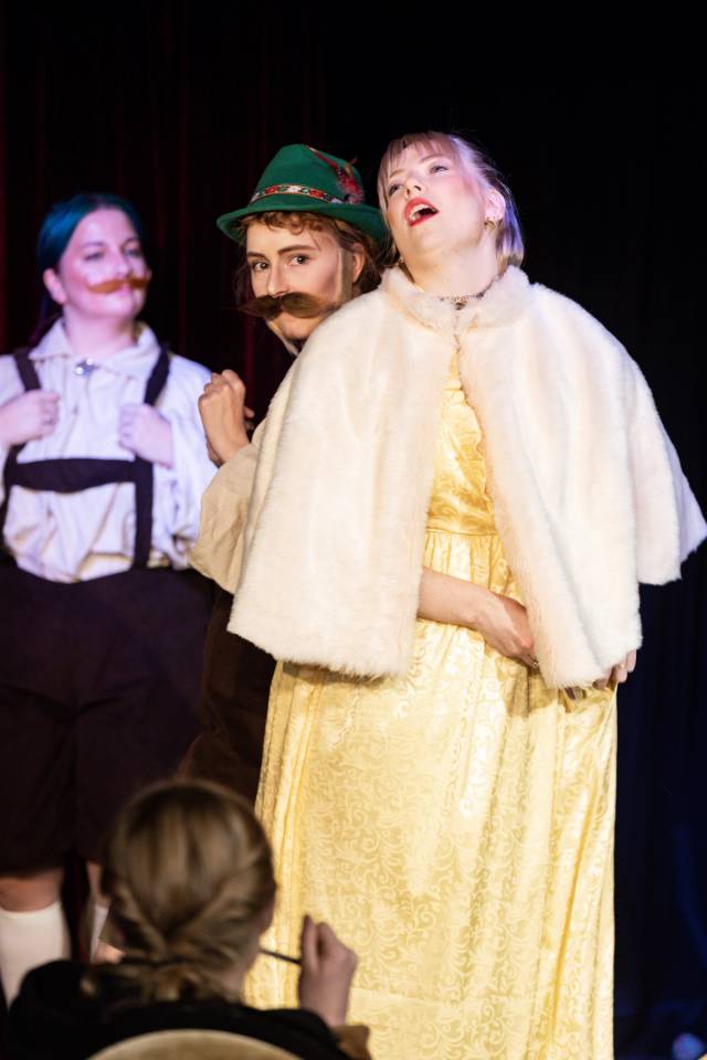 Foran står ei dame i gul kjole og ser ut som hun stønner av lykke. Over skulderen hennes ser vi en mann med løsbart og grønn hatt. I bakgrunnen står en tredje mann i lederhosen og ser på dem.