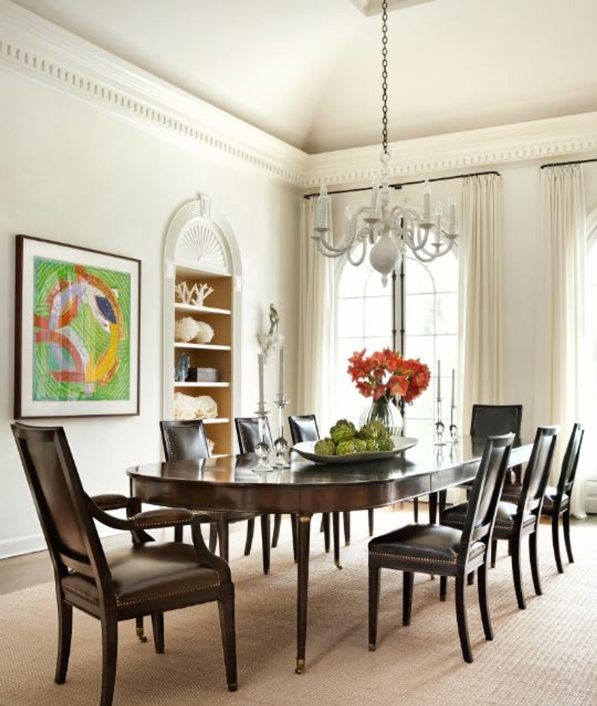 Une table de salle à manger ancienne richement polie et une chaise de salle à manger ébonisée avec un revêtement en cuir noir offrent un contraste graphique avec la salle à manger adjacente, baignée de blanc, avec un lustre en plâtre blanc.