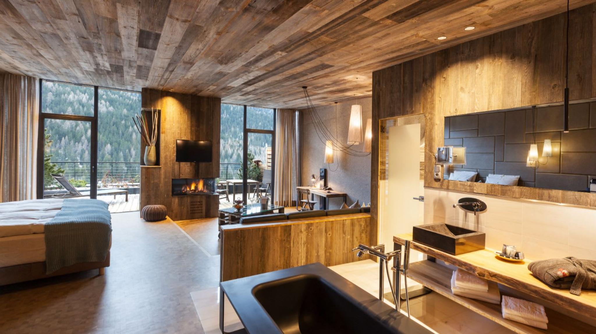 Das exquisite 'Natura Loft' mit Holzböden und einem Kamin in der Wohnzimmerecke sowie einer freistehenden Badewanne aus Lasa-Marmor mit Blick auf die große Terrasse und die offene Aussicht auf San Cassiano