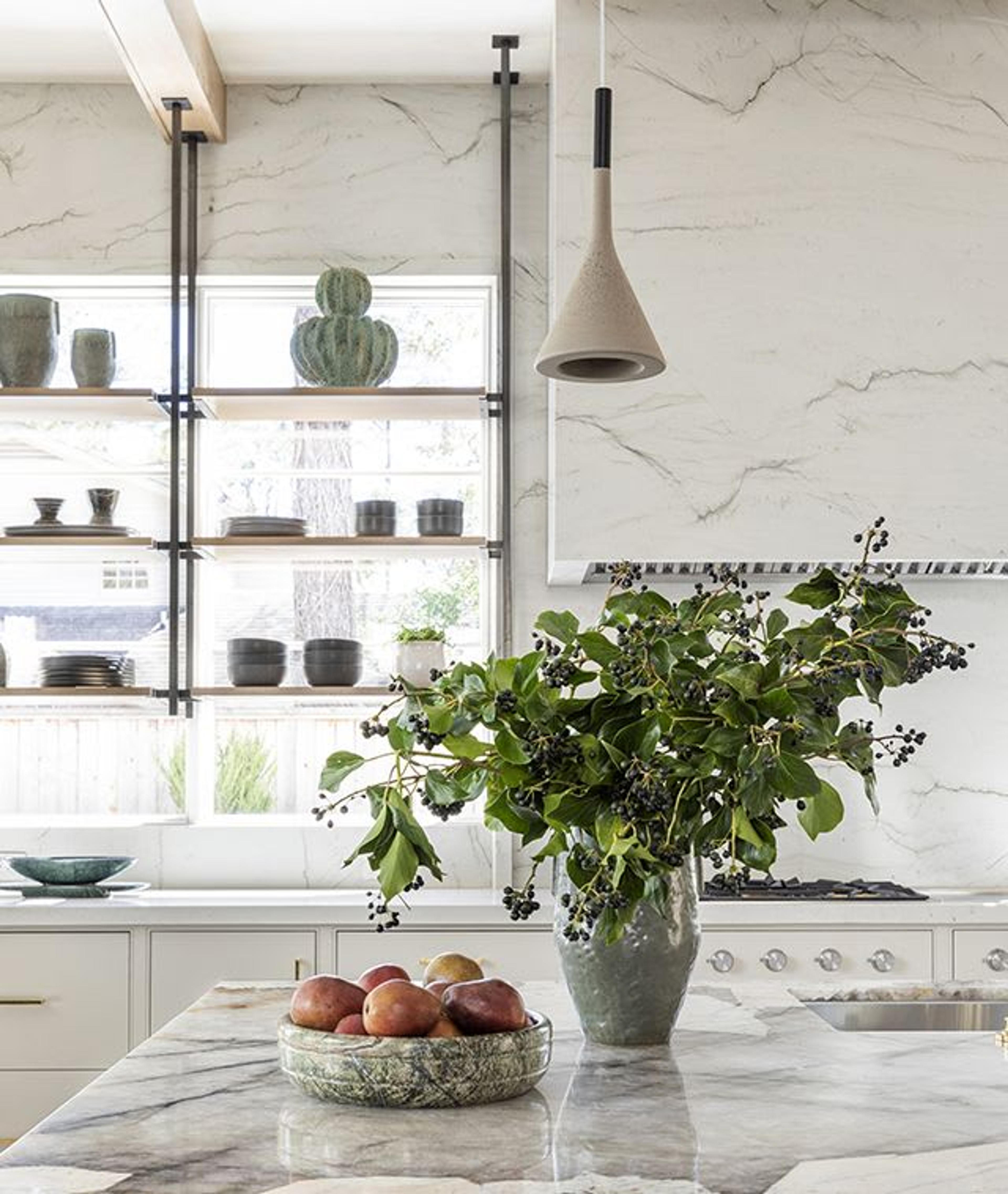 In this gorgeous European-style kitchen, suspended mixed-material open shelving serves as the centerpiece of the room while allowing ample natural light to enter the room. Quartzite stone countertops surround Gaggenau appliances and provide durable work surfaces. Minimalist concrete and blackened steel pendants provide task lighting without overpowering the space and the warm, wooden ceiling beams tie in brass details found throughout.