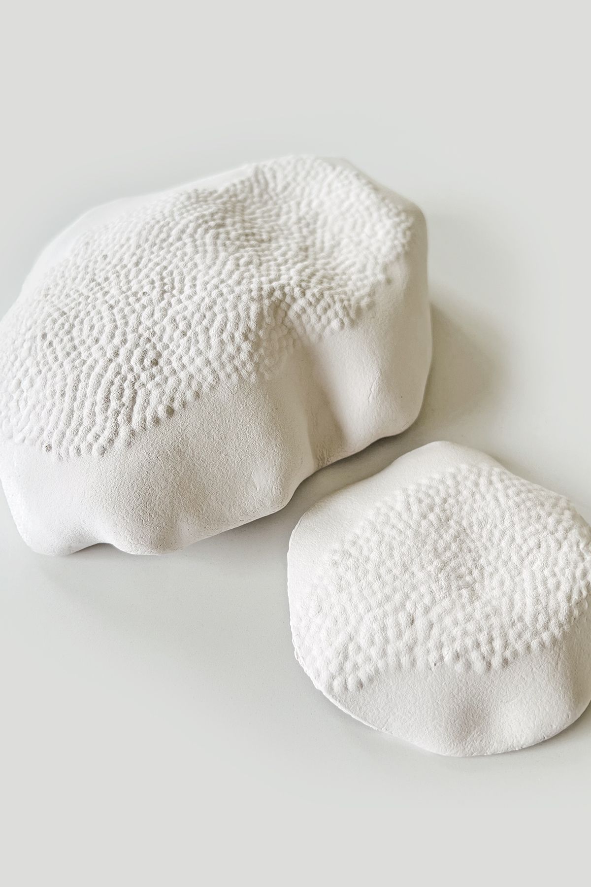 Deux sculptures en porcelaine blanche posées sur une surface claire, la plus grande marquée d'un motif de dentelle en creux.