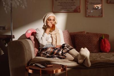 A young woman in a hijab sits comfortably on a couch, drinking from a glass with her feet propped up.