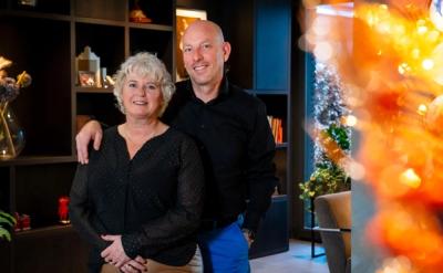 A smiling man with his arm around a woman, standing indoors with warm blurred lights in the background.