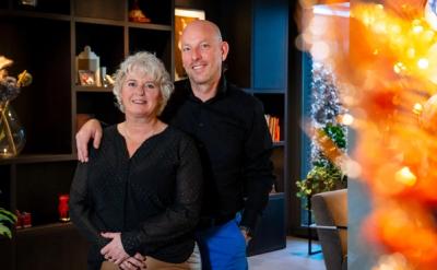 A smiling man with his arm around a woman, standing indoors with warm blurred lights in the background.