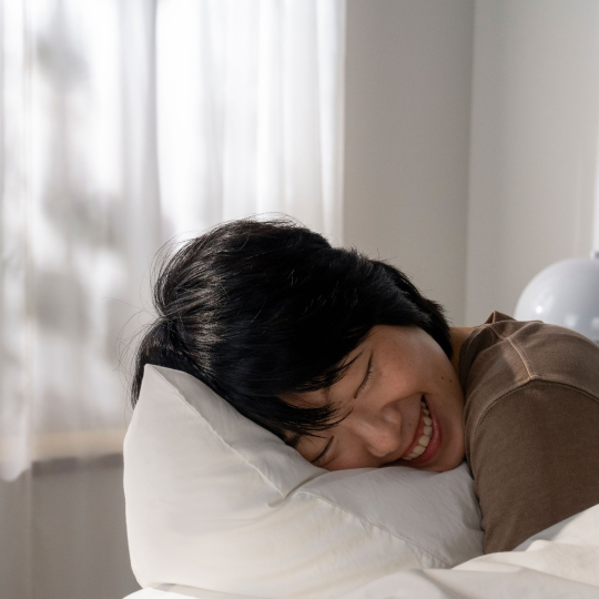 A person with dark hair smiles with closed eyes while lying on a white pillow.