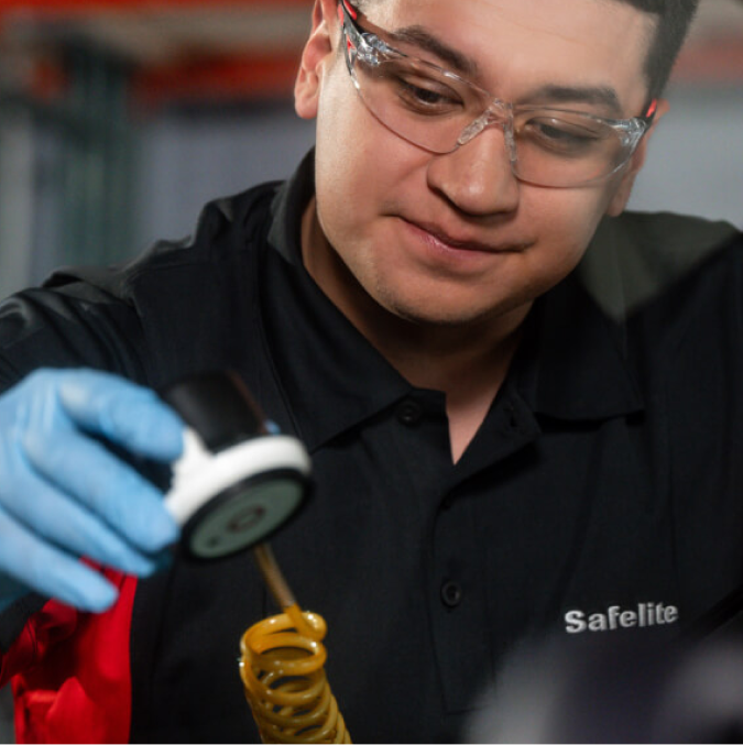 A Safelite technician in safety glasses and gloves dispenses a coiled yellow adhesive from a tool.A