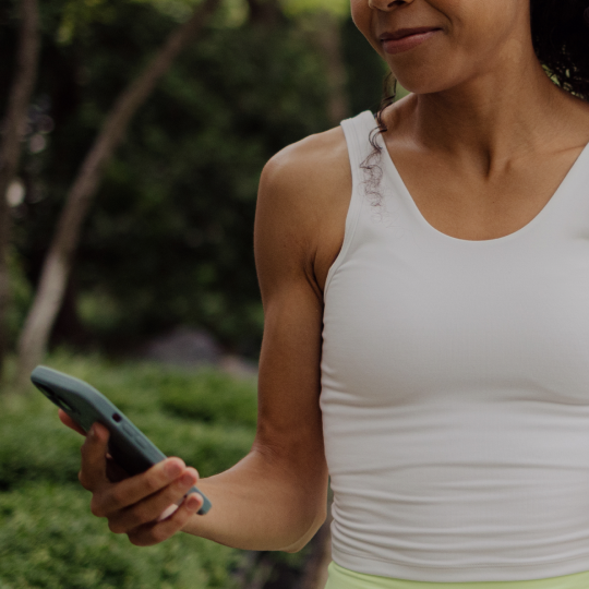 A woman in a white tank top looks at her smartphone outdoors.