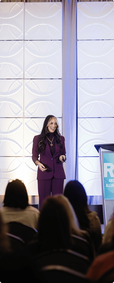 A speaker in a plum suit smiles while addressing the ATAC audience from the stage.