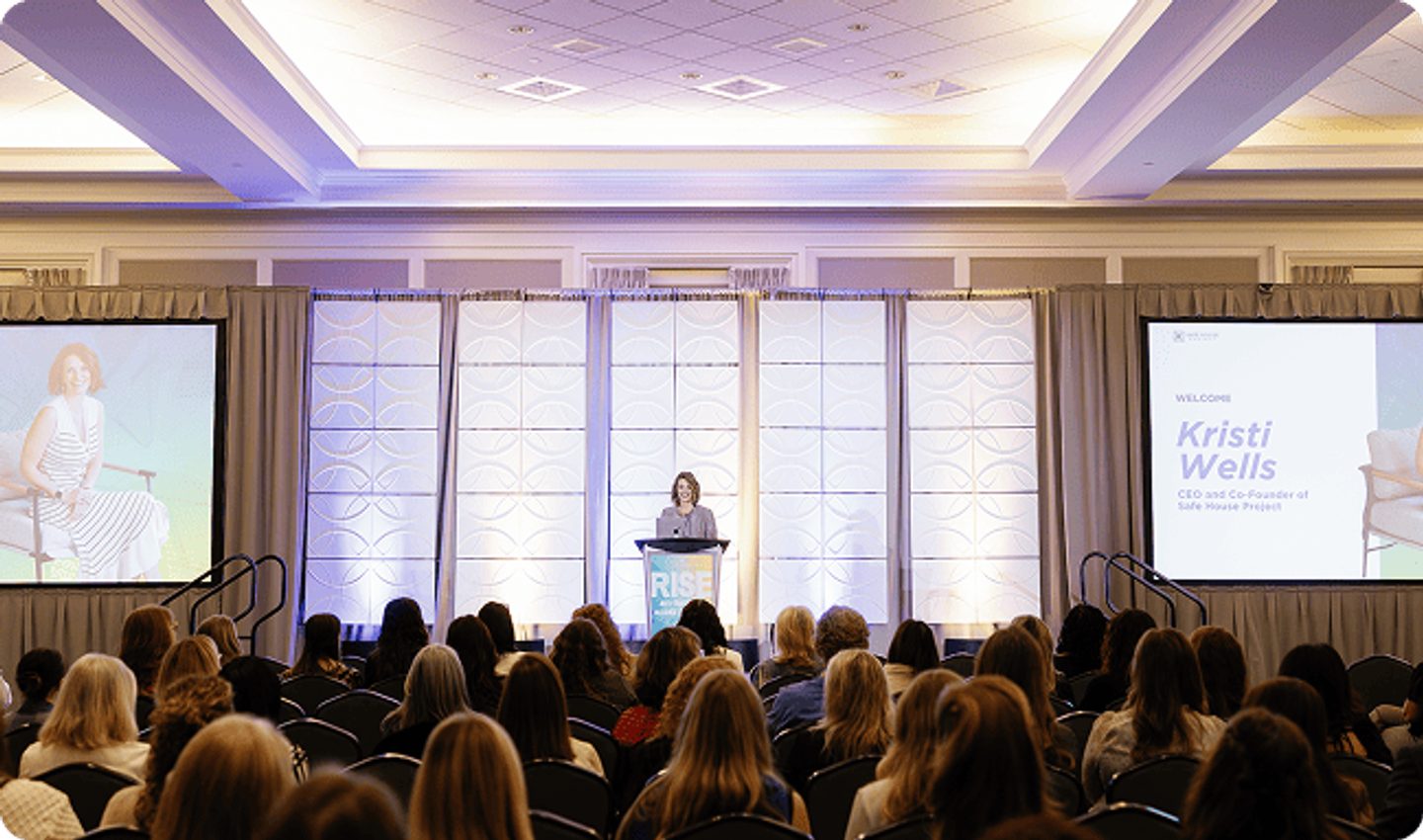 A speaker at the podium on the ATAC main stage, flanked by two screens, in front of a seated audience.
