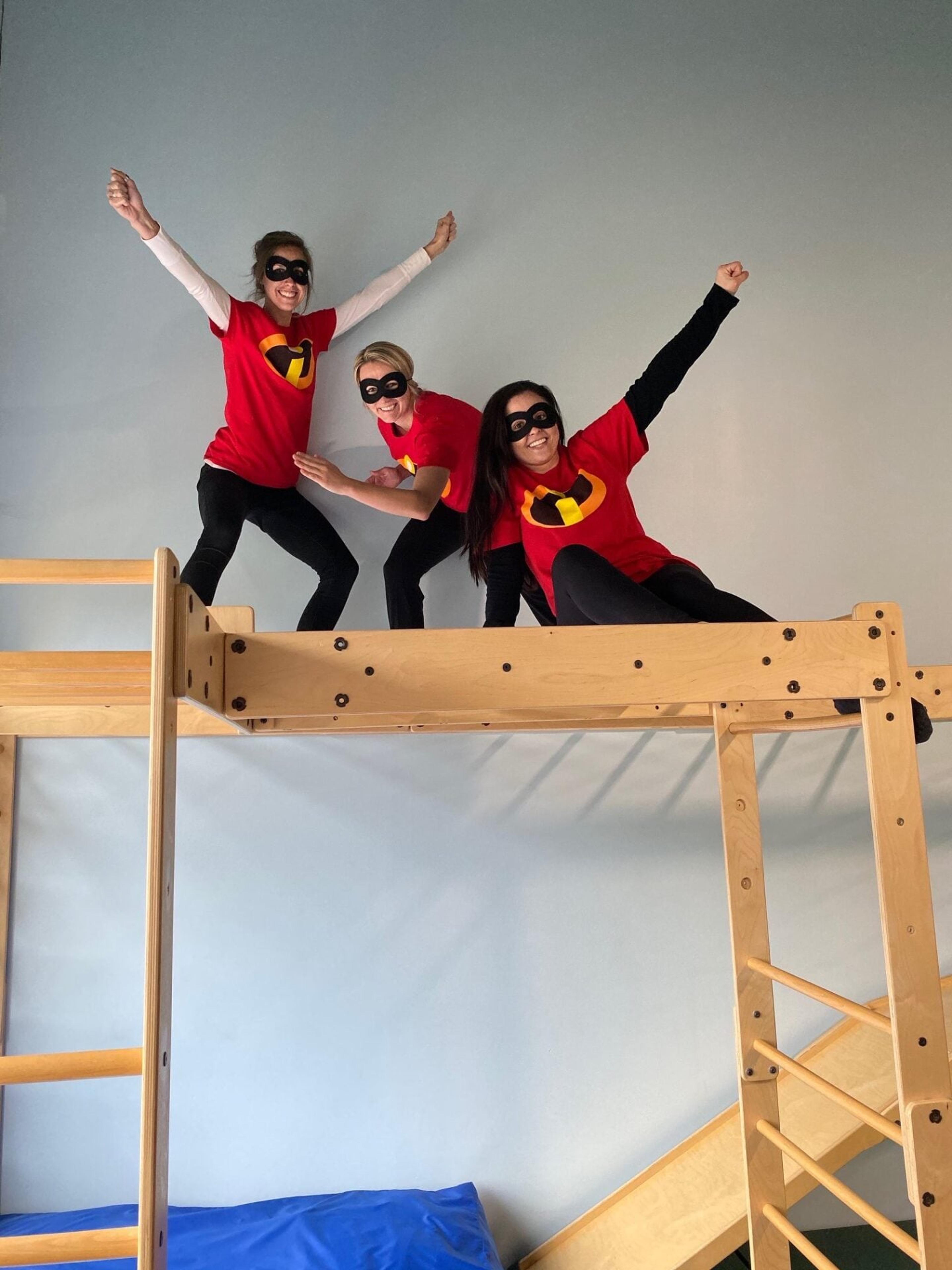 Three staff members in superhero costumes and masks strike poses on top of a wooden loft in the therapy gym
