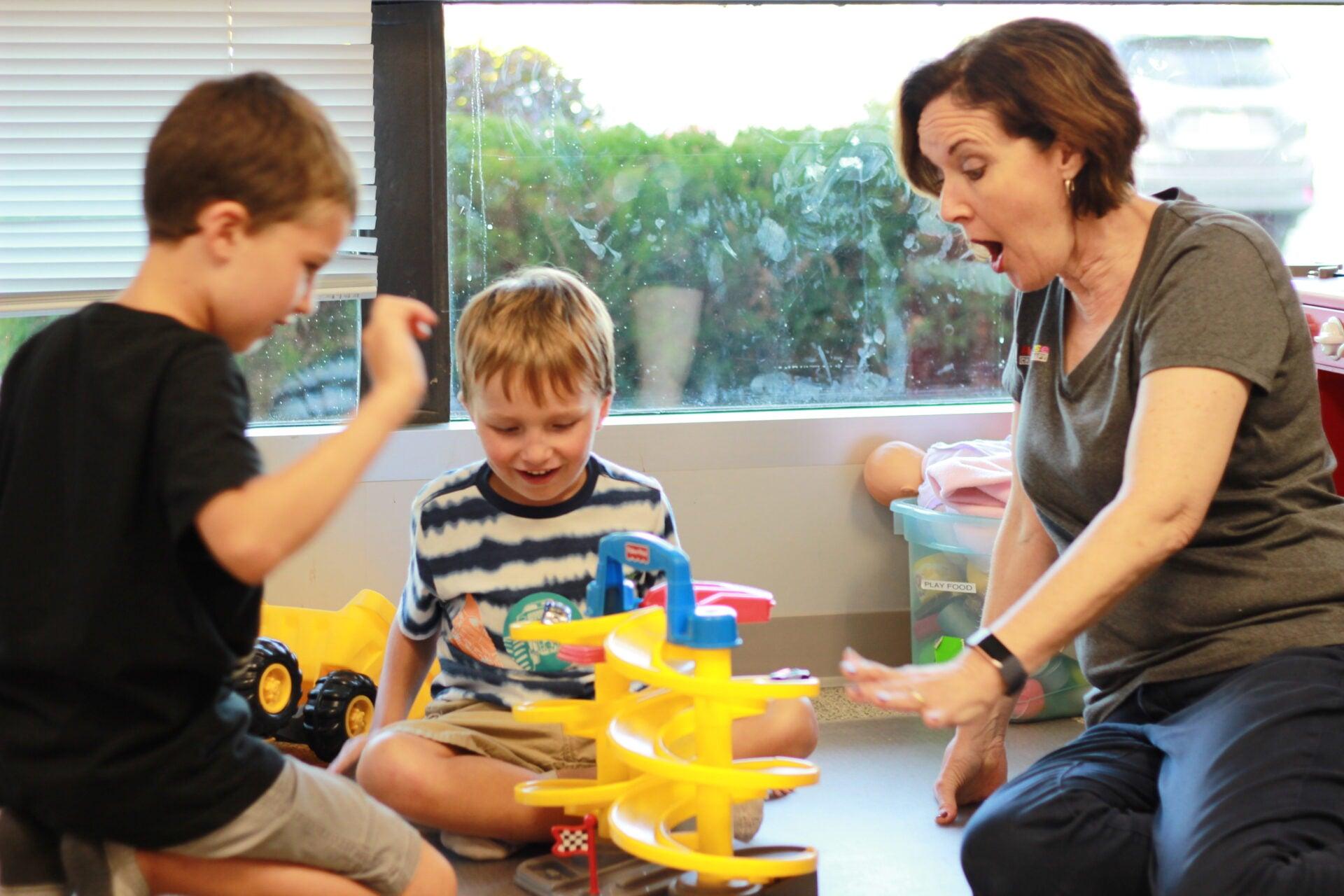 A therapist reacts with delight as two young boys send toy cars down a spiral ramp