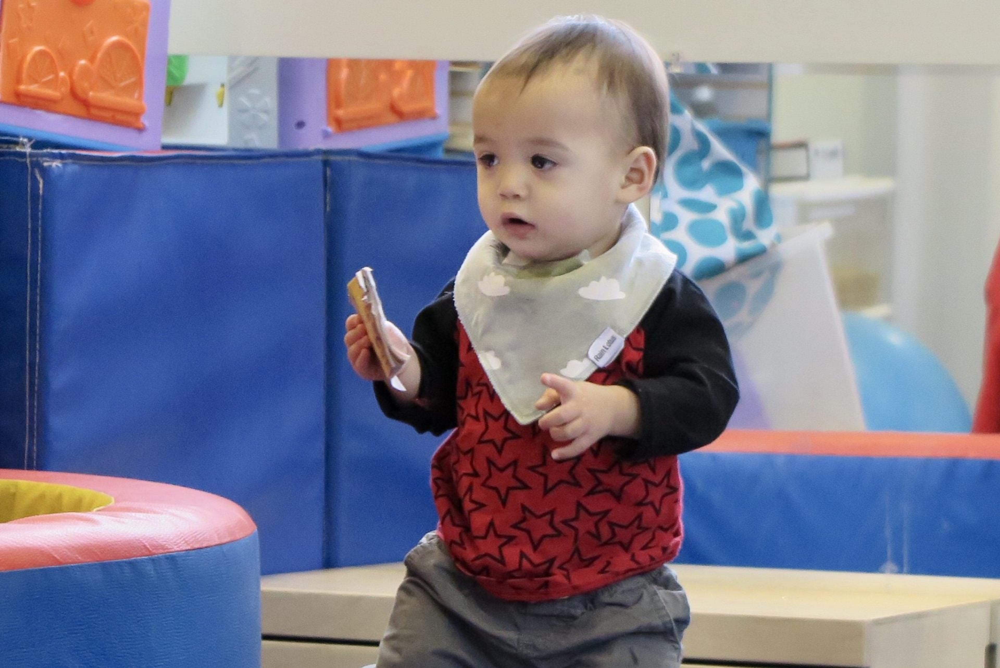 A toddler in a bandana bib stands holding a snack in a soft play area