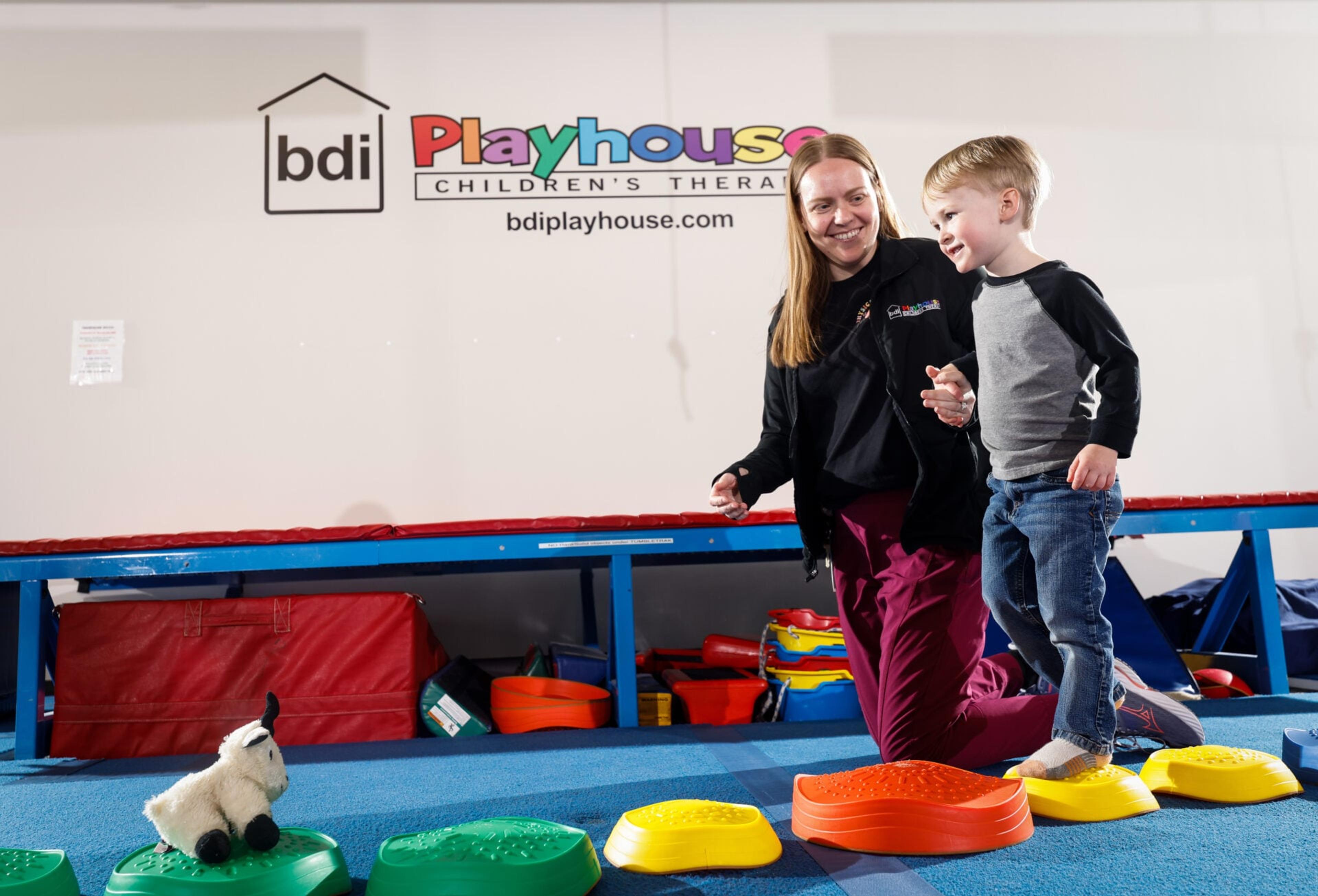 A therapist holds a boy's hand as he steps across colorful balance stones in the gym beneath the BDI Playhouse sign