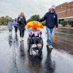 A child rides in a wheelchair under a yellow duck-themed canopy while an adult in a cap walks alongside in the rain