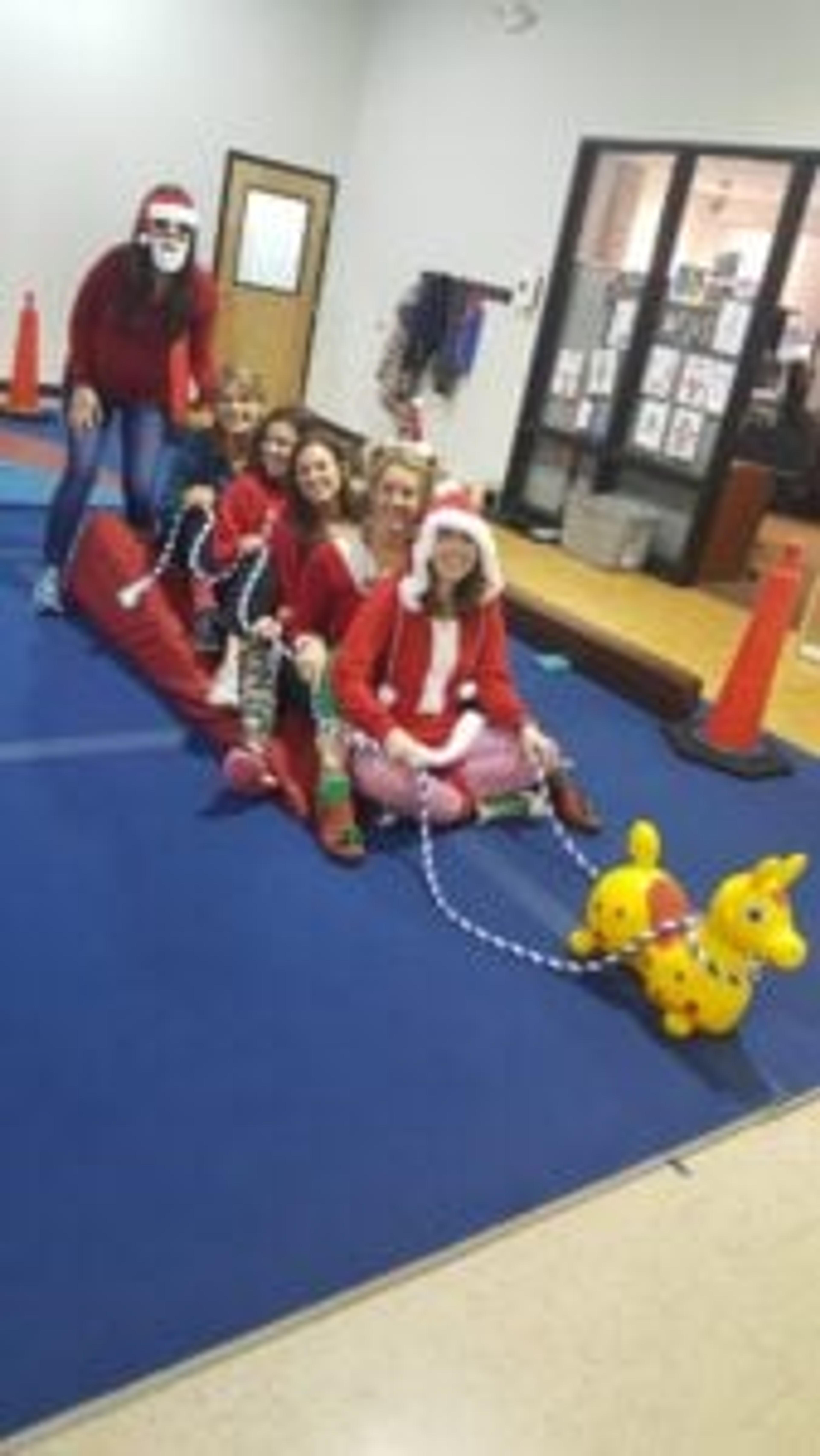 Staff in Santa hats sit in a row on the gym floor, posed like a sleigh pulled by an inflatable toy animal