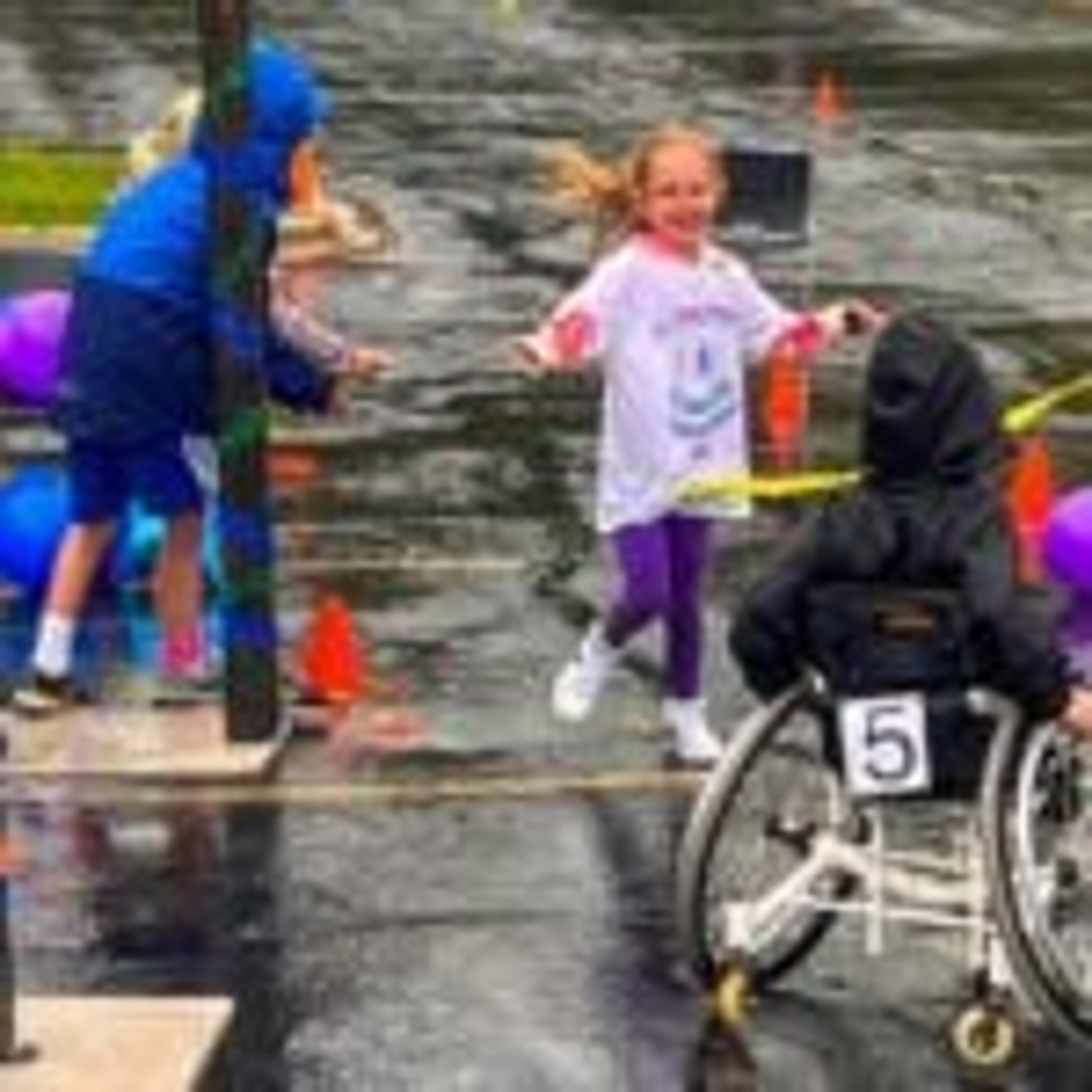 A smiling girl runs to the finish ribbon alongside a participant in a wheelchair wearing race number 5
