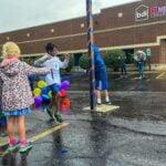 Children run past balloons and a decorated pole on a wet parking lot outside BDI Playhouse